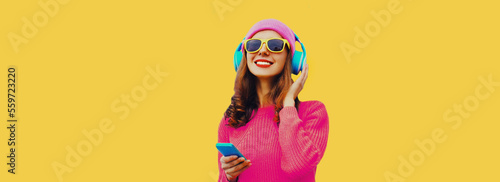 Portrait of happy smiling modern young woman in wireless headphones listening to music with smartphone wearing knitted sweater, pink hat on yellow background