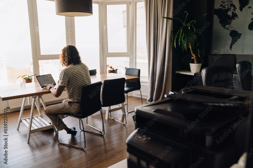 Fototapeta premium Young man using laptop and earphones while sitting by table in office