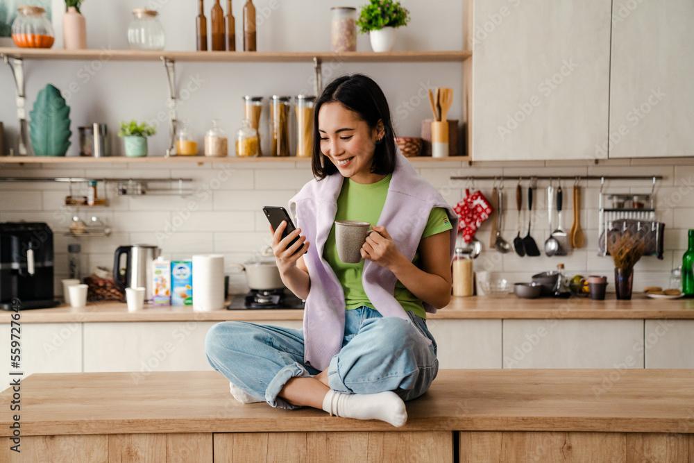 © Drobot Dean - Smiling asian woman using mobile phone and drinking coffee while sitting in kitchen © Drobot Dean - Smiling asian woman using mobile phone and drinking coffee while sitting in kitchen