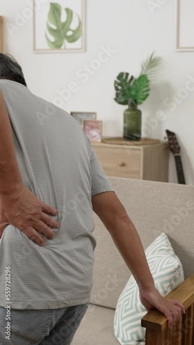 Vertical Screen: Elderly male patients slow moving and sitting on couch. Asian senior man having back pain and walking by sofa while hands holding chair arms.