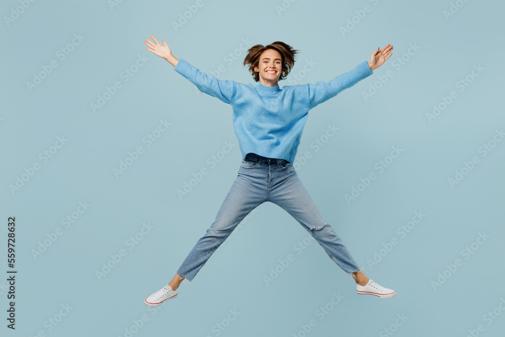 Full body overjoyed young woman wear knitted sweater look camera jump high with outstretched legs hands isolated on plain pastel light blue cyan background studio portrait. People lifestyle concept.