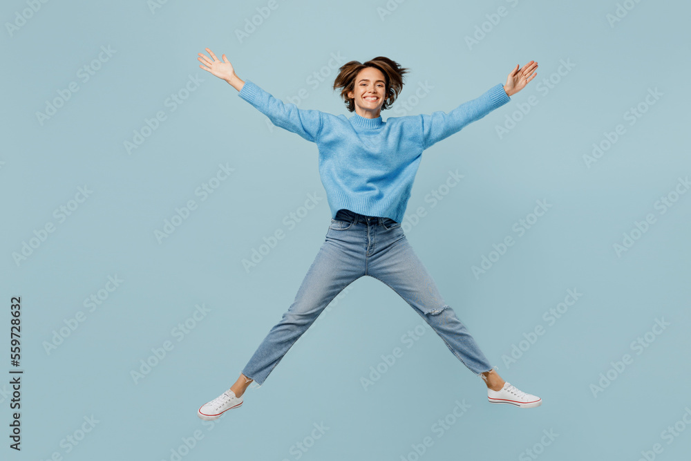 Full body overjoyed young woman wear knitted sweater look camera jump high with outstretched legs hands isolated on plain pastel light blue cyan background studio portrait. People lifestyle concept. Full body overjoyed young woman wear knitted sweater look camera jump high with outstretched legs hands isolated on plain pastel light blue cyan background studio portrait. People lifestyle concept.