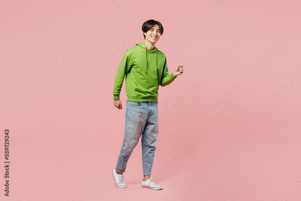 Full body sideways smiling young man of Asian ethnicity wear green hoody look camera walking going strolling isolated on plain pastel light pink background studio portrait. People lifestyle concept. Full body sideways smiling young man of Asian ethnicity wear green hoody look camera walking going strolling isolated on plain pastel light pink background studio portrait. People lifestyle concept.