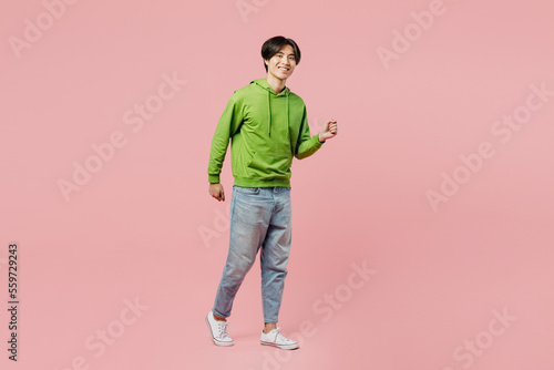 Full body sideways smiling young man of Asian ethnicity wear green hoody look camera walking going strolling isolated on plain pastel light pink background studio portrait. People lifestyle concept.