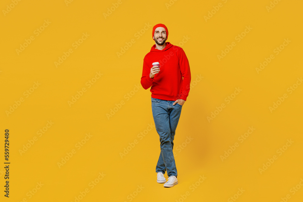 Full body young happy man wear red hoody hat look camera hold takeaway delivery craft paper brown cup coffee to go isolated on plain yellow color background studio portrait. People lifestyle concept.