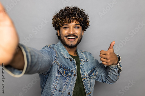 Photography Young smiling happy fun Indian man doing selfie shot pov on mobile cell phone isolated on plain grey background studio portrait