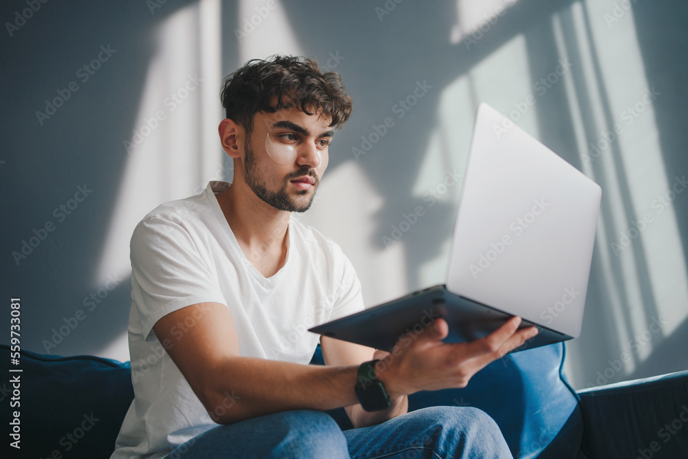 Young caucasian man wearing eye patches working online from home using a laptop. Apartment interior. Digital leisure. Young caucasian man wearing eye patches working online from home using a laptop. Apartment interior. Digital leisure.