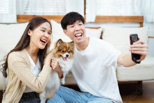 Happy cheerful Asian young couple taking a selfie photography or making a selfie video call on smartphone with their little dog in living room. Man and woman smiling to camera on smartphone. © DG PhotoStock
