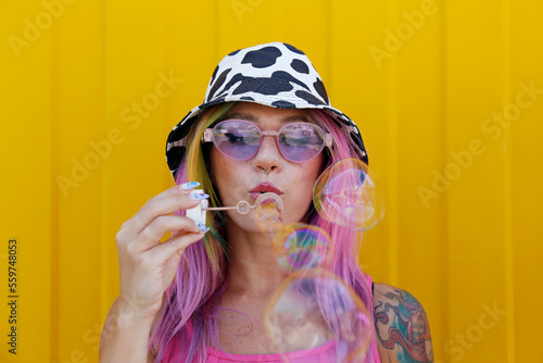 Young woman wearing hat blowing soap bubbles in front of yellow wall photo