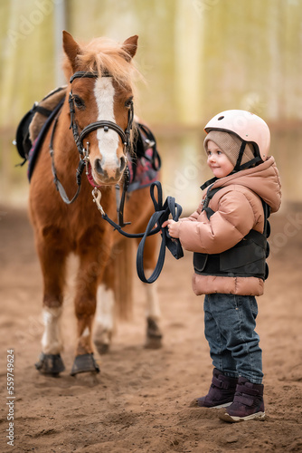 Portrait of little girl in protective jacket and helmet with her brown pony before riding Lesson © dtatiana