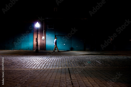 A young man walks beneath a street light in a cobblestone alley. photo