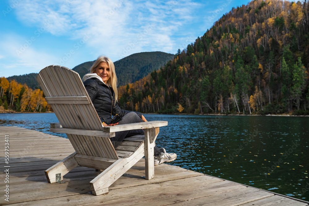 Fototapeta premium Woman resting in Comfortable wooden sun lounger Adirondack, Westport against of colorful mountains with emerald lake