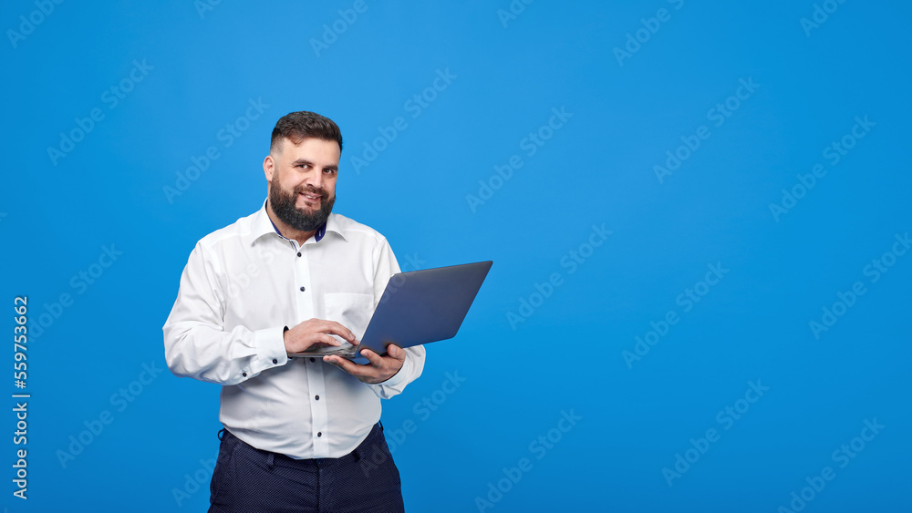 A bearded man holds a laptop in his hands on a blue background. A solid man in a white shirt works at a laptop and looks into the camera.