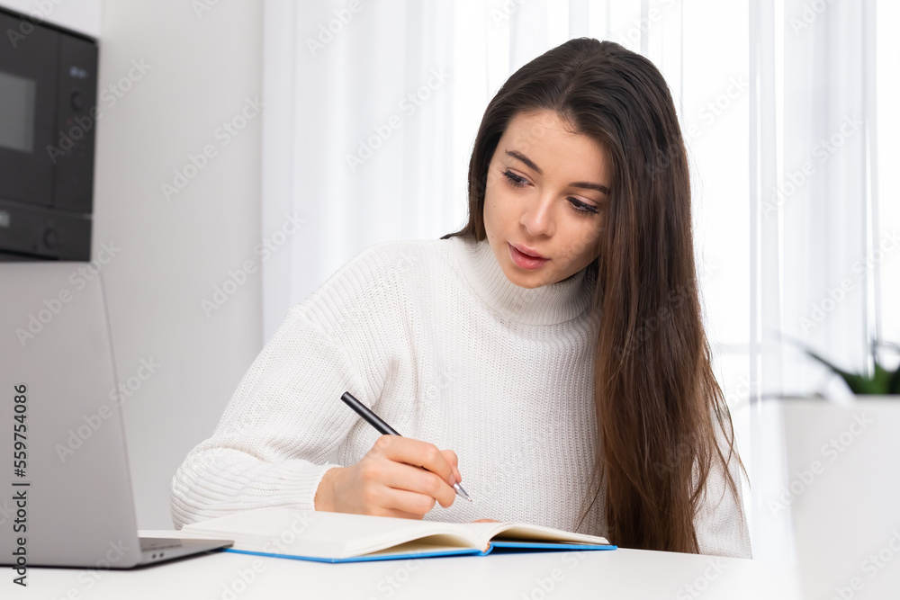 Focused female student makes notes in notepad while listening to lecture via Notebook. Young woman gets online education studying from home Focused female student makes notes in notepad while listening to lecture via Notebook. Young woman gets online education studying from home