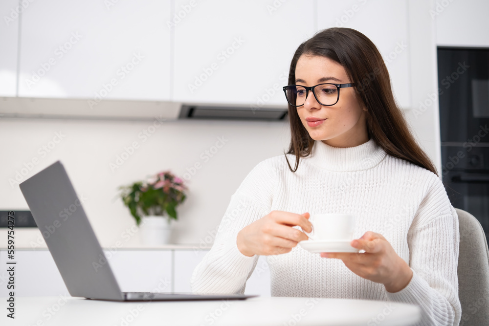 Confident young woman with long dark hair and glasses sitting at table with opened laptop and drinking coffee. SMM manager working at home. Confident young woman with long dark hair and glasses sitting at table with opened laptop and drinking coffee. SMM manager working at home.