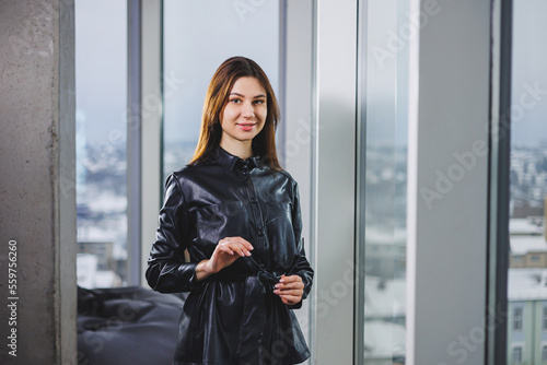 Portrait of a young slender woman in glasses and a black leather shirt. Modern woman on the background of the window in the office with a large window