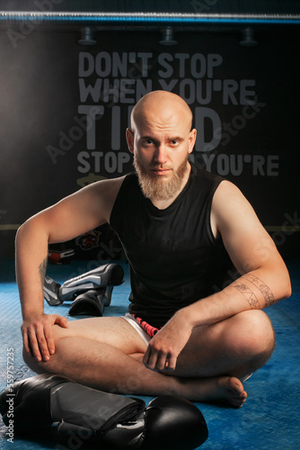 bald man with beard sitting in the gym behind boxing gloves. looking at the camera