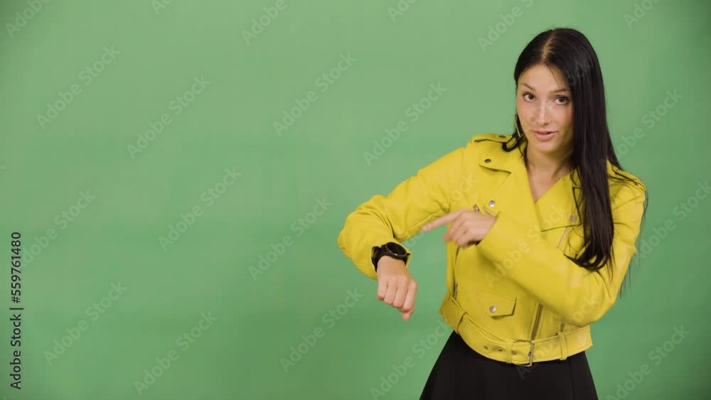 A young beautiful Caucasian woman taps her watch and looks at the camera - green screen studio A young beautiful Caucasian woman taps her watch and looks at the camera - green screen studio