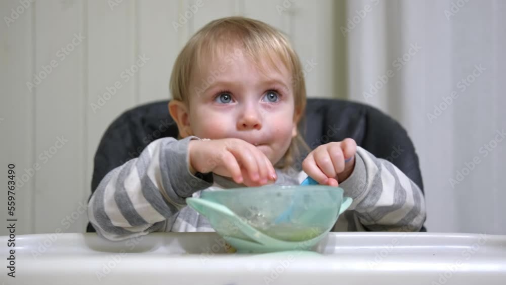 One year old hungry baby girl in striped casual clothes sits at white table in a gray highchair and eats herself with spoon. Blurred dining room background. Healthy eating for kids. Child's nutrition. One year old hungry baby girl in striped casual clothes sits at white table in a gray highchair and eats herself with spoon. Blurred dining room background. Healthy eating for kids. Child's nutrition.