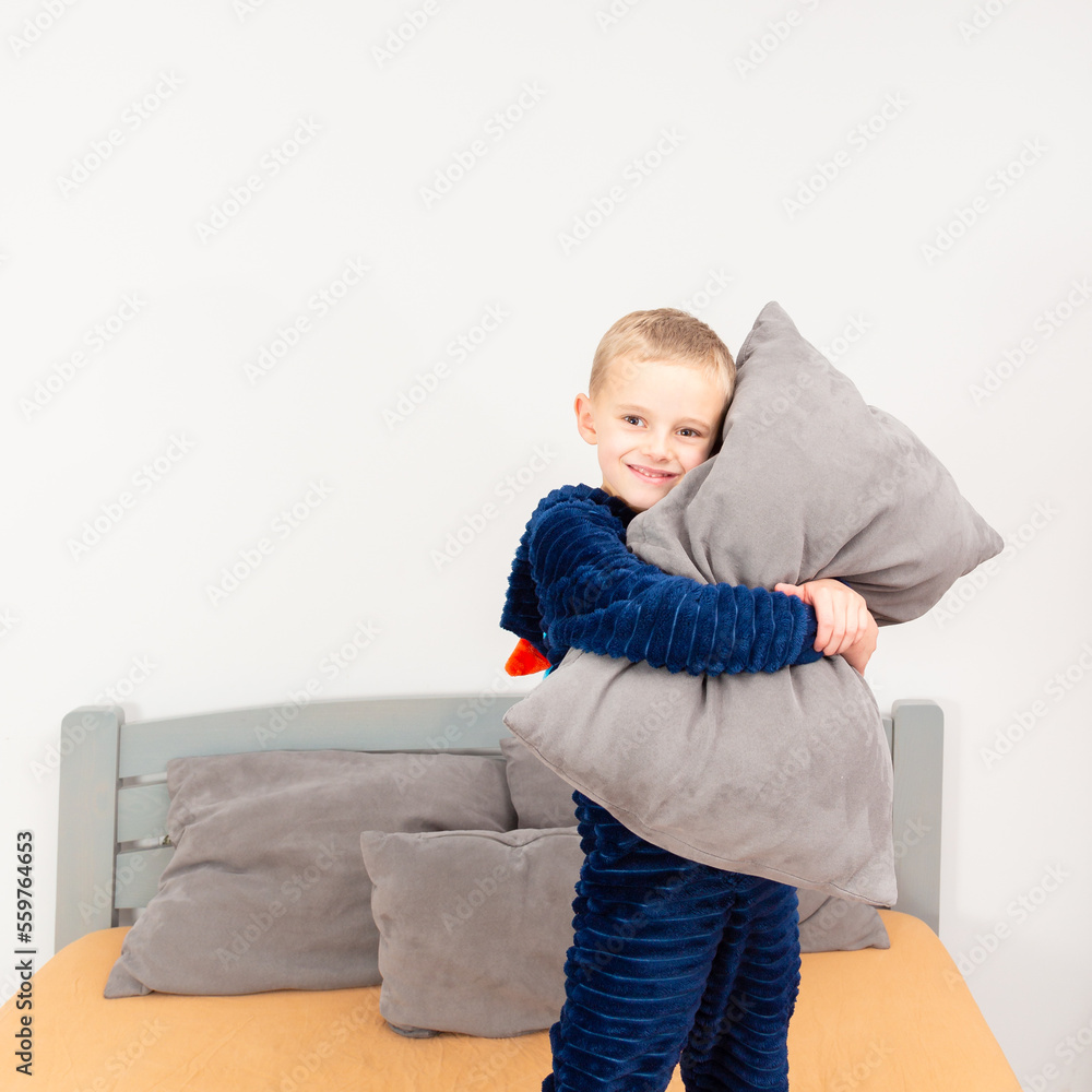 A six-year-old boy of European appearance is dressed in blue pajamas. The boy is waving and hugging with a gray pillow. A six-year-old boy of European appearance is dressed in blue pajamas. The boy is waving and hugging with a gray pillow.