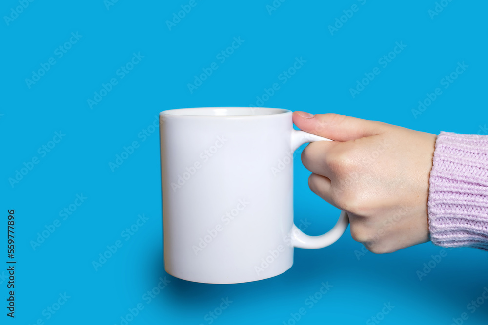 Obraz premium A hand holds a white cup with a hot drink on a blue background. The hand of a woman dressed in a sweater holds a large ceramic cup against a bright blue background. Concept of drinking hot drinks