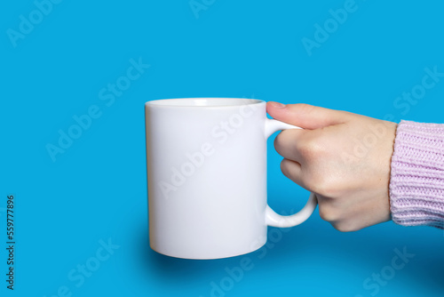 A hand holds a white cup with a hot drink on a blue background. The hand of a woman dressed in a sweater holds a large ceramic cup against a bright blue background. Concept of drinking hot drinks