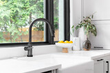 Kitchen sink detail shot in a modern, renovated kitchen with black window frames, a dark faucet, white cabinets, farmhouse sink, and cozy decor.	