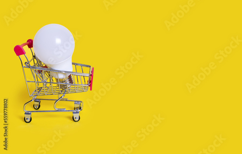 A small shopping cart and an energy-saving light bulb on a yellow background. Favorable purchase of electric power equipment to save costs. Sale of electrical equipment. Free space for text