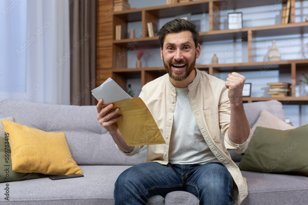 Portrait of happy man at home sitting on sofa rejoicing and celebrating victory and success looking at camera and holding hand up gesture of triumph, man received envelope mail letter with good news.