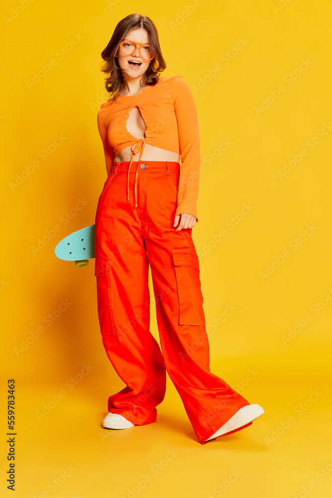 Excited girl in orange croptop and trousers with skateboard over colorful yellow background. Youth, fashion, actvie lifestyle, happy emotions and active lifestyle concept Excited girl in orange croptop and trousers with skateboard over colorful yellow background. Youth, fashion, actvie lifestyle, happy emotions and active lifestyle concept