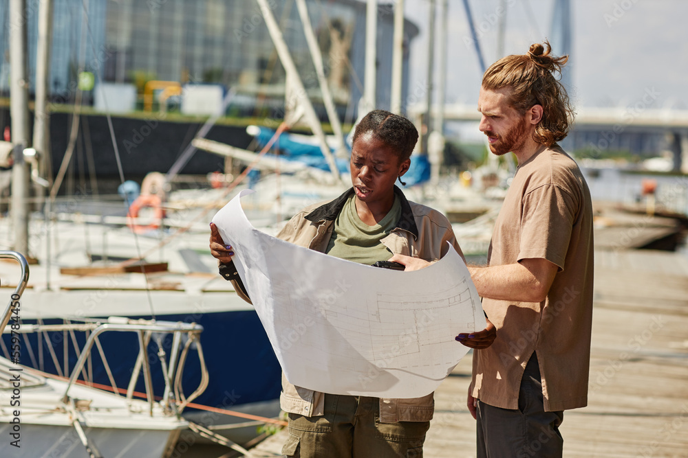Waist up portrait of two workers looking at blueprints in yacht docks lit by sunlight, copy space