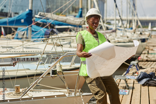 Portrait of black young woman looking at blueprints while working in yacht docks lit by sunlight, copy space