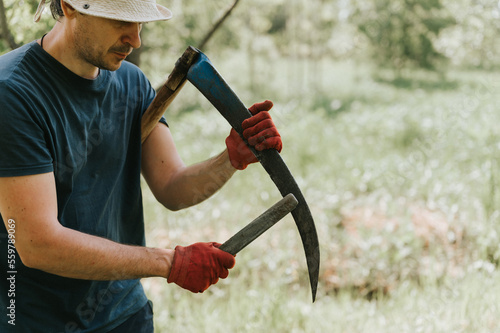 mowing grass traditional old-fashioned way with hand scythe on household village farm. young mature farmer man sharpening the scythe with grass or whetstone for mowing the grown weed of a farmland photo