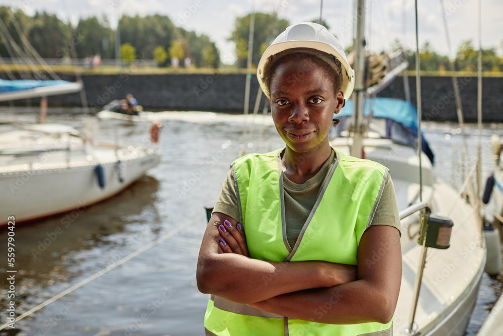 Waist up portrait of black young woman wearing hardhat while standing in yacht docks outdoors, copy space