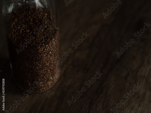 close-up of a bottle filled with flax seed, large quantity of flax seed in a glass container. Linseed reserve on wood table. 