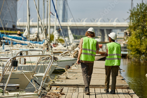 Full length back view of two workers wearing hardhats while walking on pier, copy space