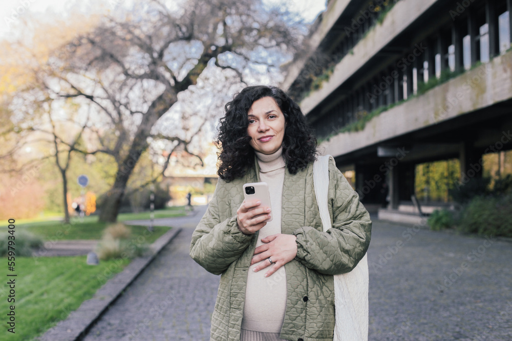 A pregnant woman touching her belly, holding a pink smartphone in an urban park A pregnant woman touching her belly, holding a pink smartphone in an urban park