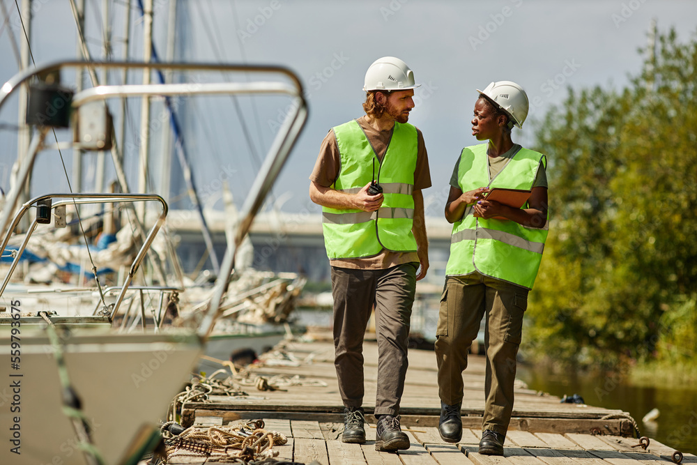 Full length portrait of two young workers wearing hardhats while walking towards camera on pier, copy space Full length portrait of two young workers wearing hardhats while walking towards camera on pier, copy space