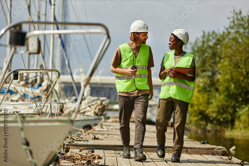 Full length portrait of two young workers wearing hardhats while walking towards camera on pier, copy space photo