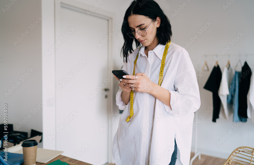 Happy sewer using smartphone while standing near table in store Happy sewer using smartphone while standing near table in store
