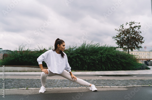 Young woman makes deep side lunges to pump the muscles of the legs and buttocks, simple and effective exercises during morning jog around city. Healthy lifestyle and keeping the body in shape © BullRun