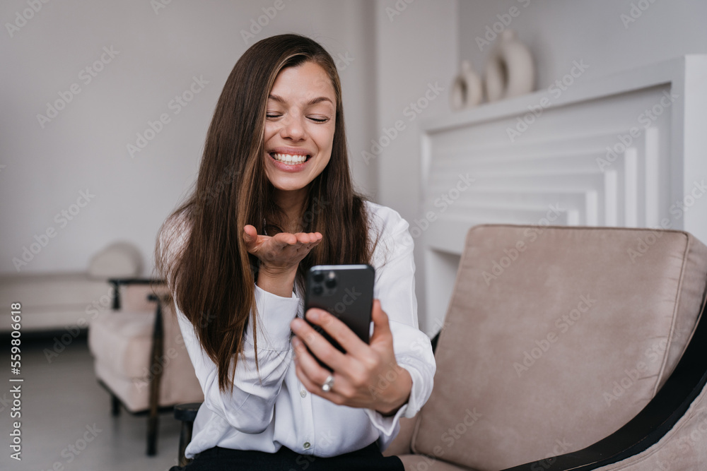 Naklejka premium Cheerful Hispanic young woman holds phone sends air kiss during video call sitting on cozy chair at home dressed in white shirt. Pretty brunette businesswoman grateful to her husband. Happy woman.