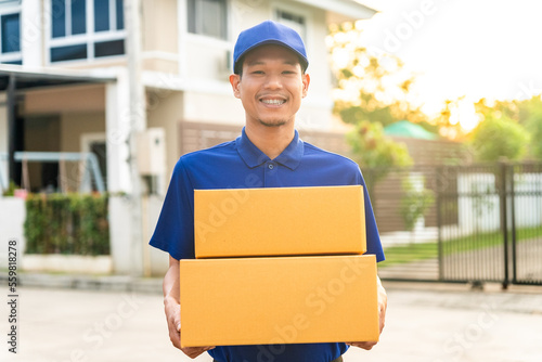 Handsome asian smiley delivery man wearing blue uniform holding parcel cardboard boxes in front of customer house and he so happy with his job. © BJ Day Stock