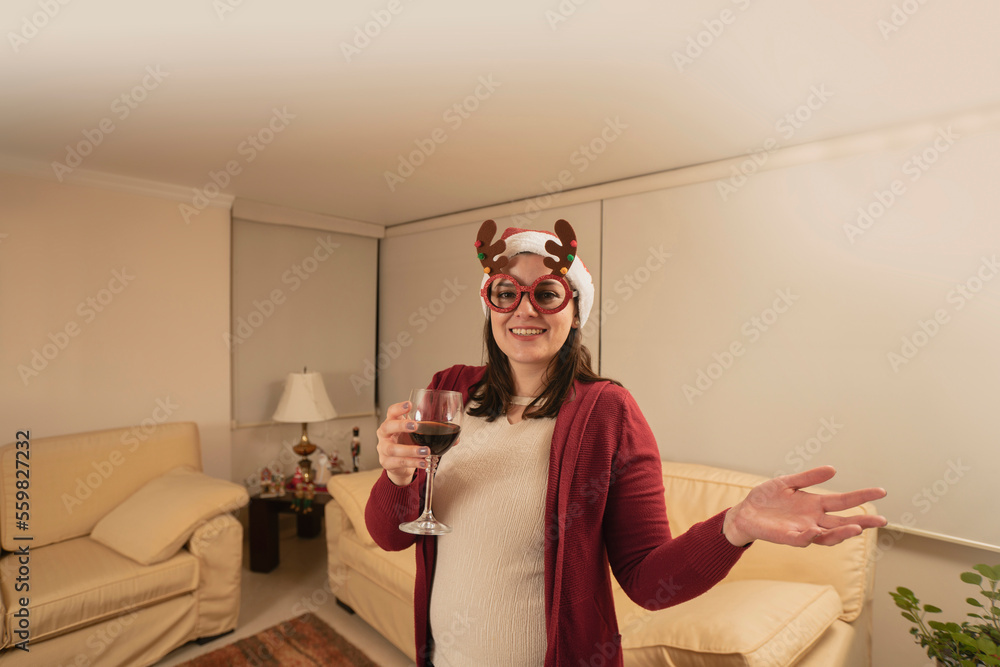 woman dressed up with reindeer glasses and christmas hat standing looking at the camera, toasting with a glass of red wine in her living room at night