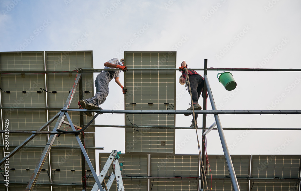 View from below of two men solar technicians standing on metal rail and mounting photovoltaic solar panel. Male workers installing solar modules under white blue sky. Concept of sustainable energy.