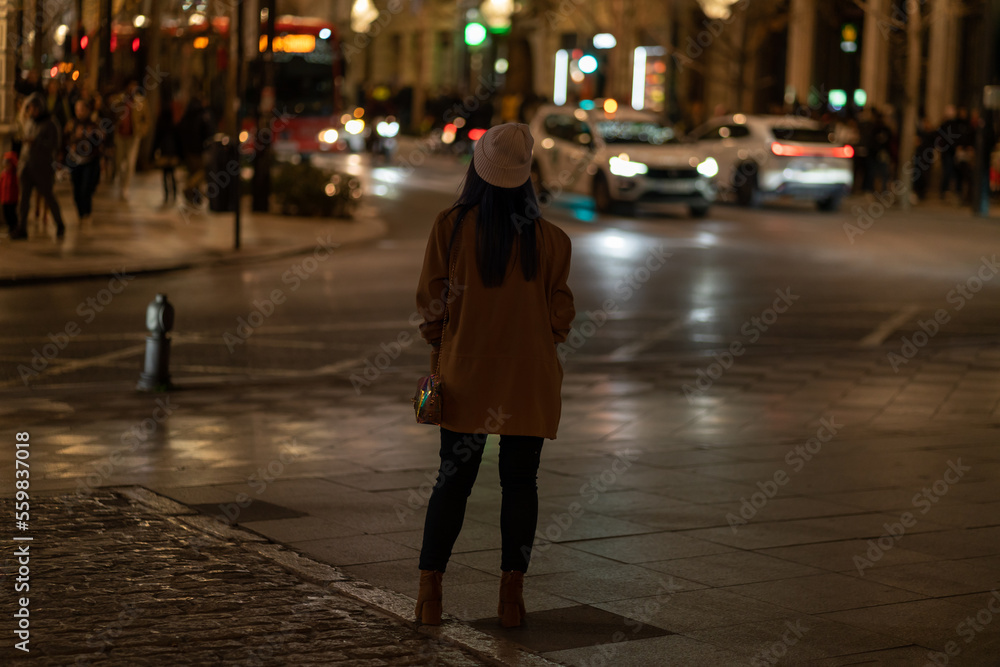Rear view of young woman looking at night city lights, wearing brown jacket and hat.