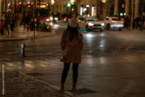 Rear view of young woman looking at night city lights, wearing brown jacket and hat.