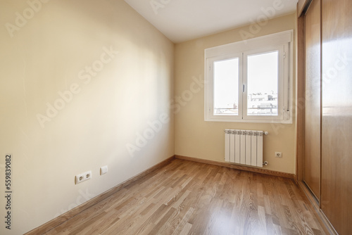 Room with a built-in wardrobe with three sliding oak doors with skirting boards and matching floors and smooth cream-colored walls and a white aluminum window with two hinged leaves