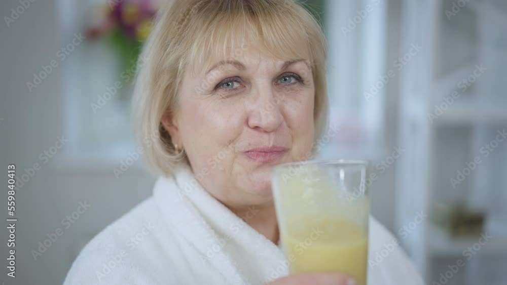 Close-up beautiful senior woman drinking healthy vitamin smoothie in slow motion looking at camera. Smiling confident Caucasian retiree enjoying tasty healthful drink at home in the morning posing Close-up beautiful senior woman drinking healthy vitamin smoothie in slow motion looking at camera. Smiling confident Caucasian retiree enjoying tasty healthful drink at home in the morning posing