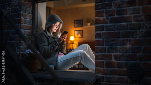 White Woman Wearing a Hoodie Using her Smartphone While Sitting on Windowsill at Night. Female Gamer Playing Addictive Mobile Phone Games Instead of Sleeping. Bad Case of Unhealthy Gaming Addiction